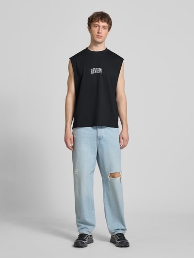 Männermodel trägt Muskelshirt mit Logo und Rundhalsausschnitt von REVIEW in Schwarz, Baumwolle, Regular Fit, Ganzkörperansicht von vorne.