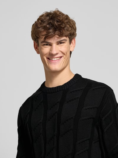Mannenmodel draagt Regular Fit Strickpullover aus reiner Baumwolle van Tommy Jeans, zwart, kabelpatroon, puur katoen, crew neck, logo detail, lange mouw, ribboorden, unie kleur, detailopname bovenlichaam.