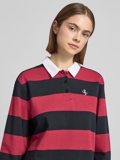 Frauenmodel trägt Polo-Langarmshirt mit Streifen und kontrastiger Knopfleiste von Review aus reiner Baumwolle im Regular-Fit mit gesticktem Logo in Kontrastfarbe, horizontale breite Streifen in Rot und Schwarz, weißer Polo-Kragen, Nahaufnahme Oberkörper.