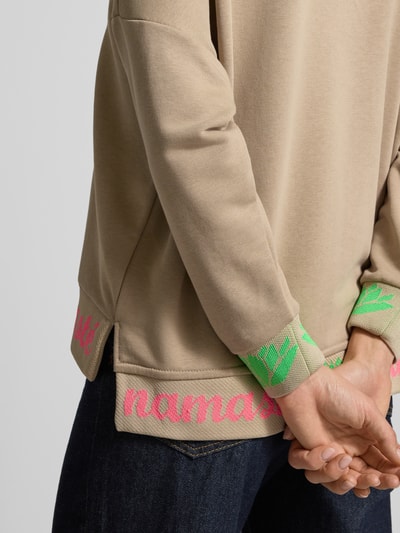 Männermodel trägt Oversized Hoodie mit Strasssteinbesatz Modell 'Namaste' von miss goodlife in Beige, Statement-Print, Detailaufnahme Rückansicht.