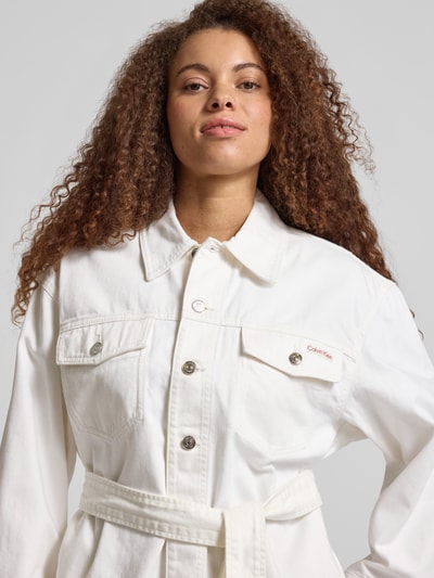 Frauenmodel trägt Regular Fit Jeansjacke mit abnehmbarem Taillenband von Calvin Klein Jeans in Weiß, reine Baumwolle, Nahaufnahme Oberkörper. Brusttaschen, Umlegekragen, Logo-Patch und Gürtel betonen Struktur und Details.