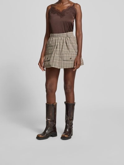 Frauenmodel trägt Minirock mit Pattentaschen von BAUM & PFERDGARTEN im karierten Material-Mix mit Baumwoll-Anteil, Falten-Optik, Nahtreißverschluss, ausgestellte Passform, Farbtöne Braun und Beige, Bundweite bei Größe 36: 36 cm, Ganzkörperansicht von vorne.