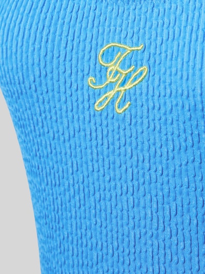 Frauenmodel trägt Badeanzug mit Logo-Stitching von TOMMY HILFIGER in kräftigem Blau, Crinkle-Optik, gelbes Logo, Nahaufnahme Oberkörper.
