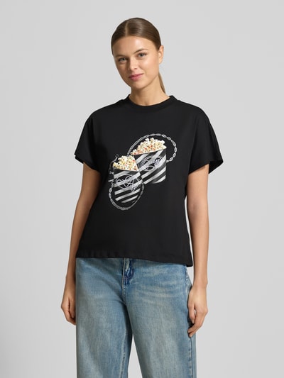 Kobiece modelka nosi Regular Fit T-Shirt mit Ziersteinbesatz Modell 'Popcorn' od Guess, czarna bawełna, motyw print, okrągły dekolt, przód, zdjęcie całej sylwetki.