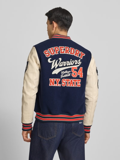 Męskmodel nosi College-Jacke mit Patches und Eingrifftaschen von Superdry, mieszanka bawełny, krój regular fit, stójka, listwa guzikowa, wpuszczane kieszenie, długość tyłu w rozmiarze M: 65 cm, szerokość ramion w rozmiarze M: 45 cm, szerokość klatki piersiowej w rozmiarze M: 49 cm, długość rękawów w rozmiarze M: 70 cm, widok całej sylwetki od tyłu