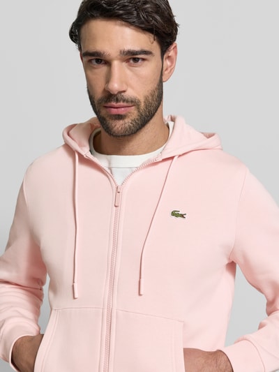 Männermodel trägt Regular Fit Sweatjacke aus Baumwoll-Mix von Lacoste in Rosa mit Logo-Patch, Kapuze und Reißverschluss, Nahaufnahme Oberkörper.
