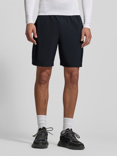 Männermodel trägt schwarze Straight Fit Shorts mit Cargo-Taschen, Modell 'Vibe' von Under Armour, elastischer Bund, Ganzkörperansicht von vorne.