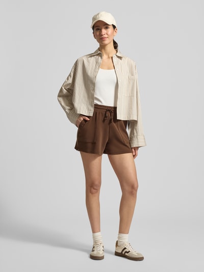 Frauenmodel trägt Sweatshorts mit Seitentaschen von Scotch & Soda in Braun, gerader Schnitt, elastischer Bund, Ganzkörperansicht von vorne.