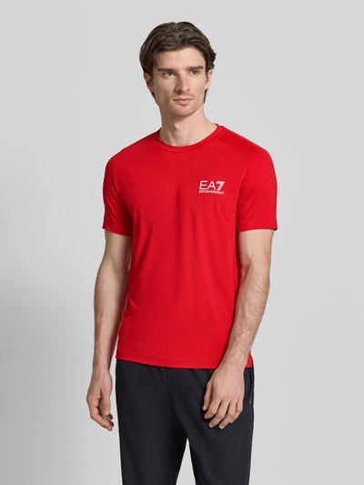 Männermodel trägt T-Shirt mit Label-Print von EA7 Emporio Armani in Rot, Slim Fit, glatte Textur, Nahaufnahme Oberkörper.