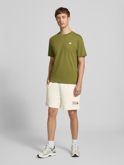 Männermodel trägt Sweatshorts aus Baumwoll-Mix von New Balance in Creme mit Label-Print, elastischem Bund und Gesäßtasche, Ganzkörperansicht von vorne.