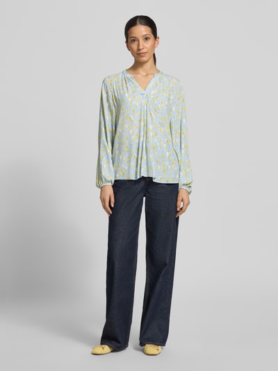 Frauenmodel trägt Regular Fit Bluse aus reiner Viskose mit Allover-Motiv-Print von Emily Van den Bergh in Hellblau mit Gelb, Tunika-Ausschnitt, langärmelig, elastische Ärmelabschlüsse, Rückenlänge 68 cm, Ärmellänge 65 cm, Ganzkörperansicht von vorne.