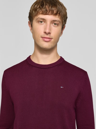 Männermodel trägt Slim Fit Pullover aus reiner Baumwolle von Tommy Jeans in unifarbenem Burgunder, Strickstruktur, Nahaufnahme Oberkörper.
