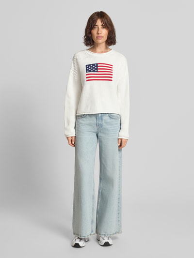 Vrouwenmodel draagt gebreide pullover met Amerikaanse vlag print van GAP, puur katoen, regular fit, grof tricot, ronde hals, ruglengte bij maat S 47 cm, mouwlengte 51 cm, lichtgrijs, rechte snit. Ganzkörperansicht van voren.