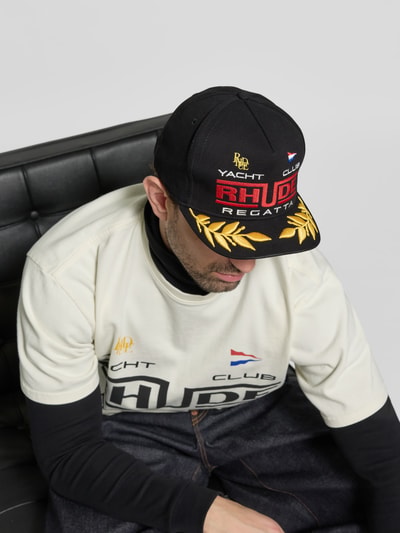 Männermodel trägt Basecap aus reiner Baumwolle von RHUDE mit verstärktem Schirm, verstellbarem Riegel, Label-Stitching, Belüftungslöcher, Nahaufnahme Oberkörper.