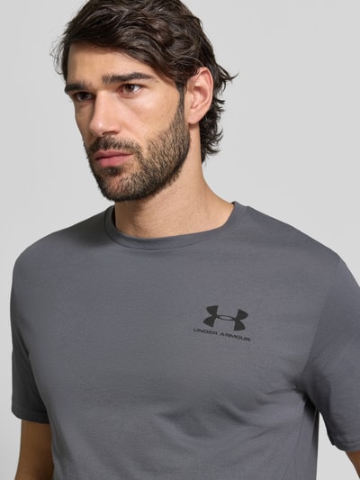 Męski model nosi Regular Fit T-Shirt mit Logo-Print Modell 'Left' von Under Armour, mieszanka bawełny, okrągły dekolt, kolor szary, nadruk z logo na piersi, długość tyłu w rozmiarze M: 72 cm, zbliżenie na górną część ciała z przodu