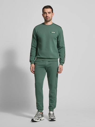 Männermodel trägt Sweatshirt mit Label-Details und Rundhalsausschnitt von EA7 Emporio Armani aus grünem Baumwoll-Mix, Regular-Fit, Ganzkörperansicht von vorne. Rückenlänge bei Größe M: 63 cm, Ärmellänge bei Größe M: 66 cm.