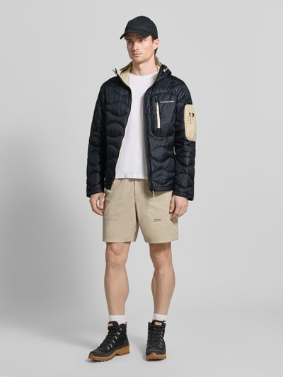 Männermodel trägt Regular Fit Jacke in Stepp-Optik mit Logo-Stitching von PEAK PERFORMANCE, schwarze und beige Farben, Ganzkörperansicht von vorne. Kontext: Herren Jacke, Kapuze mit Tunnelzug, Eingrifftaschen mit Reißverschluss, Napoleontasche, Rückenlänge 71 cm, Schulterbreite 47 cm, Brustweite 56 cm, Ärmellänge 67 cm.