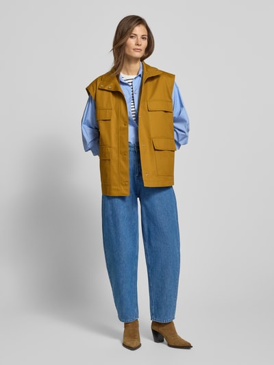 Modelka prezentuje Oversized Weste mit aufgesetzten Taschen - curated by Tinvcb od Tinvcb, kolor musztardowy, krój relaxed fit, stójka, listwa guzikowa, kieszenie z patkami, długość tyłu w rozmiarze 36: 67 cm, szerokość ramion w rozmiarze 36: 52 cm, szerokość w biuście w rozmiarze 36: 59 cm, styl casual, jeansy i botki, widok całej sylwetki z przodu.