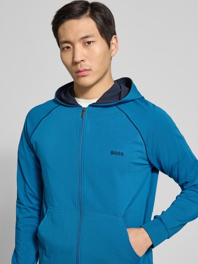 Männermodel trägt Regular Fit Sweatjacke aus Baumwoll-Mix von BOSS in Optik: Unifarben mit Reißverschluss, Label-Stitching-Applikation, Kontraststreifen, Kapuze, Langarm, Ärmelabschluss: Gerippt, Rumpfabschluss: Gerippt, Kängurutasche, Materialverarbeitung: Sweat, Farbe Blau, Nahaufnahme Oberkörper.