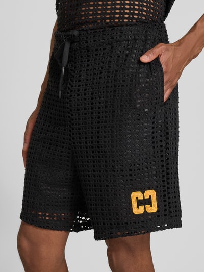 Männermodel trägt Straight Fit Shorts in Loch-Optik von COLUCCI, schwarzer elastischer Material-Mix, gelbes Logo-Stitching, Nahaufnahme Oberschenkel.