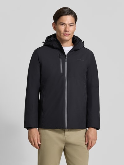 Męski model nosi Regular Fit Jacke mit Logo-Print od Lindbergh z elastycznej mieszanki materiałów, czarna, nadruk z logo, kaptur z tunelem, kieszeń napoleonka, długość tyłu w rozmiarze M: 74 cm, szerokość ramion w rozmiarze M: 46 cm, szerokość klatki piersiowej w rozmiarze M: 42 cm, długość rękawów w rozmiarze M: 62 cm, widok całej sylwetki z przodu.