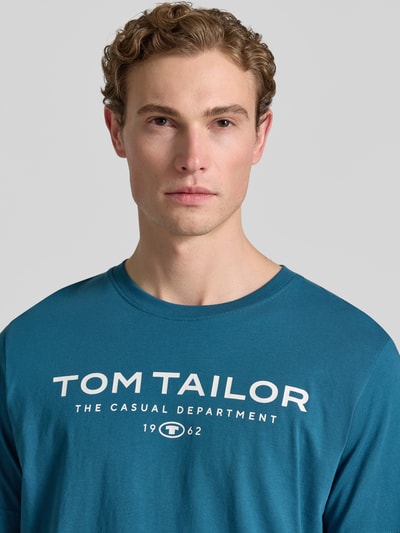 Mężczyzna nosi Regular Fit T-Shirt aus reiner Baumwolle von Tom Tailor w jednolitym kolorze z nadrukiem z logo, dżersej, czysta bawełna, krótki rękaw, dekolt okrągły, efekt: jednolity kolor, aplikacja: nadruk z logo, męski t-shirt. Nahaufnahme górnej części ciała.