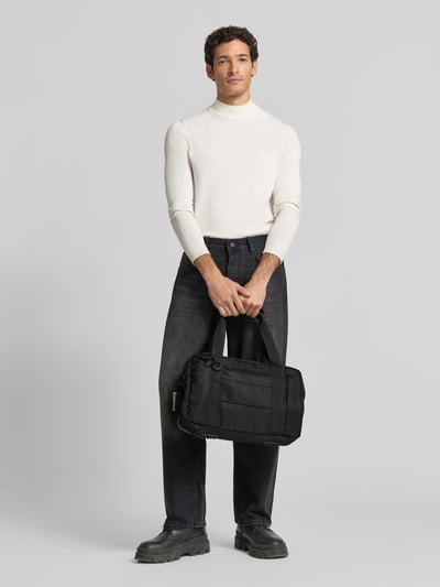 Männermodel trägt schwarzen Weekender mit Logo-Detail von AY STUDIOS, sichtbarer Reißverschluss, Netz-Taschen, Ganzkörperansicht von vorne.