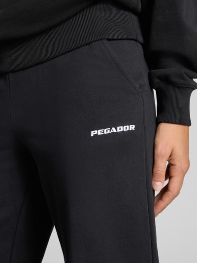 Männermodel trägt spodnie dresowe o kroju wide fit z mieszanki bawełny Model 'ANNI' od PEGADOR, detal z logo, elastyczny pas, Długość wewnętrzna nogawki w rozmiarze S: 78 cm, detailaufnahme.