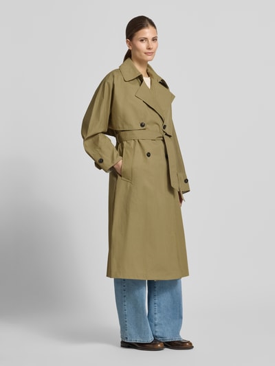 Vrouwenmodel draagt trenchcoat met verdekte sluiting en strikceintuur van Blonde No. 8, katoenmix, getailleerd model, platte kraag, zij-steekzakken, knopen, ruglengte maat 36: 113 cm, schouderbreedte: 60 cm, borstwijdte: 54 cm, mouwlengte: 49 cm, geheelopname vooraanzicht.