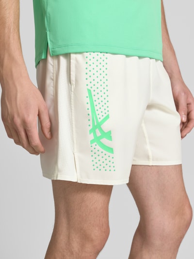 Mężczyzna nosi Regular Fit Shorts mit integriertem Slip Modell 'ICON' od ASICS TIGER, nadruk z logo, elastyczny pas, szerokość w rozmiarze M: 37 cm, długość wewnętrzna nogawki w rozmiarze M: 18 cm, kremowe z zielonym nadrukiem, gładka tekstura, zbliżenie na dolną połowę ciała