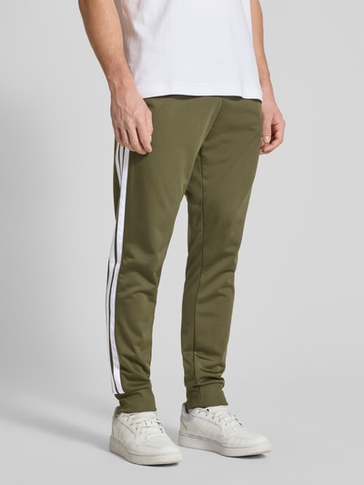 Männermodel trägt Regular Fit Sweat-Anzug mit Kontraststreifen von adidas Sportswear in Oliv mit weißen Kontraststreifen, Nahaufnahme Unterkörper.