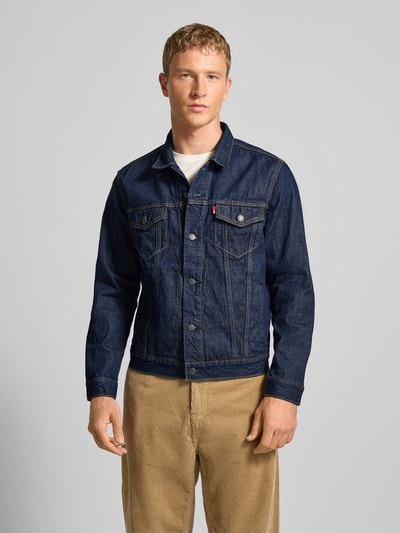 Männermodel trägt Jeansjacke mit Brusttaschen Modell 'THE TRUCKER JACKET' von Levi's®, Reine Baumwolle, Regular-Fit, Knopfleiste, Umlegekragen, Label-Detail, dunkelblaue Farbe, ganzkörperansicht von vorne.