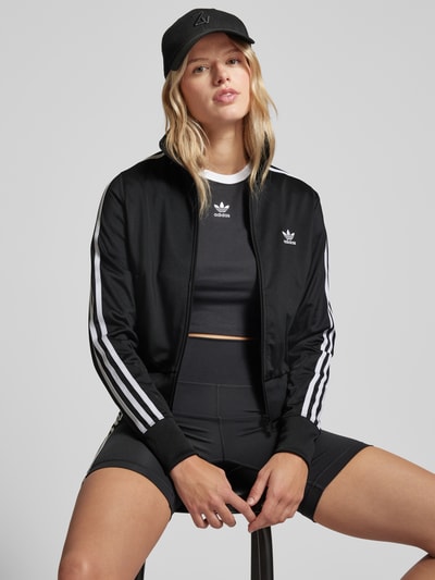 Kobiece modelka nosi czarną damską Trainingsjacke mit Logo-Stitching Modell 'FIREBIRD' od adidas Originals z białymi paskami, widok na cały korpus z przodu.