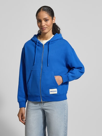 Frauenmodel trägt Sweatjacke mit Zweiwege-Reißverschluss von Smith and Soul, königsblau, Känguru-Tasche, Label-Detail, Ganzkörperansicht von vorne.