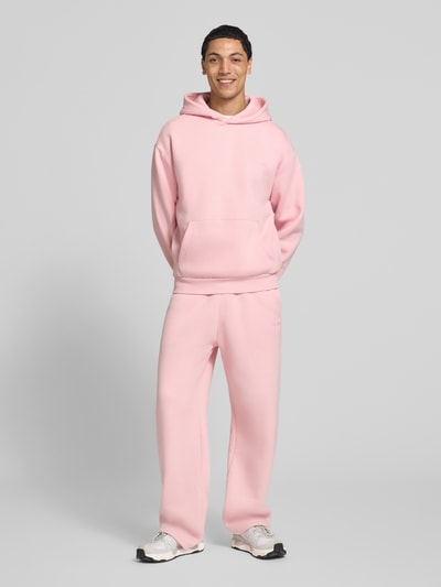 Männermodel trägt Hoodie mit Logo-Print von REVIEW in Rosa, Baumwoll-Mix, lockere Passform, Kängurutasche, Ganzkörperansicht von vorne.