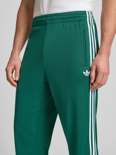Mannenmodel draagt Sweatpants mit elastischem Bund van adidas Originals in groen met witte details, losse fit, elastische taille, steekzakken, label-stitching, binnenbeenlengte maat M 81 cm, close-up van bovenlichaam.
