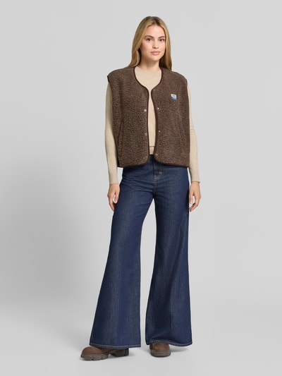 Frauenmodel trägt Relaxed Fit Weste aus Teddyfell Modell 'Hoktown' von American Vintage, braun, Knopfleiste, Logo-Patch, Ganzkörperansicht von vorne. Damen Weste, weiche Teddyfell-Textur, lockerer Schnitt, Rückenlänge 56 cm, Schulterbreite 43 cm, Brustweite 63 cm.