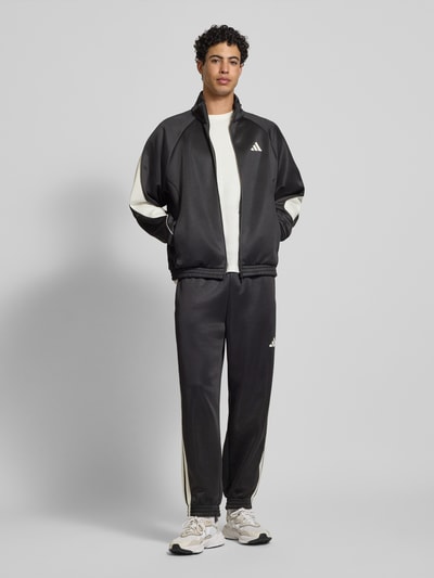 Męski model nosi Kurtka treningowa z logo i stójką od adidas Sportswear, mieszanka materiałów z bawełną, krój regular fit, długość tyłu 68 cm, długość rękawa 67 cm, całość w czerni z białymi akcentami, widok całej sylwetki z przodu.
