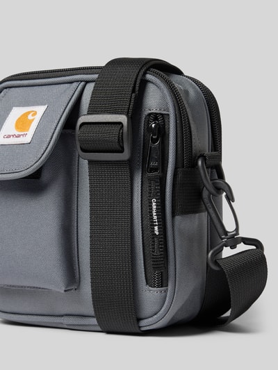 Produktfoto, Crossbody Bag mit Logo-Patch von Carhartt Work In Progress in Grau mit schwarzem, verstellbarem Schulterriemen, Detailaufnahme. Geteiltes Hauptfach mit Reißverschluss, abnehmbarer Schulterriemen, kompakte Maße für Herren.