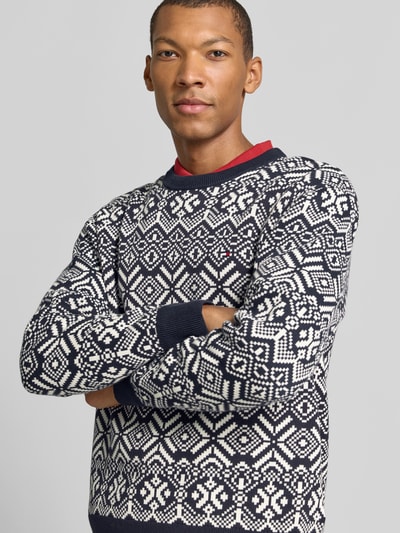 Męski model nosi Pullover mit Logo-Detail von Tommy Hilfiger, Wzór na całej powierzchni, imitacja dzianiny, okrągły dekolt, długi rękaw, prążkowane zakończenia, aplikacja: detal z logo, Ganzkörperansicht von vorne.