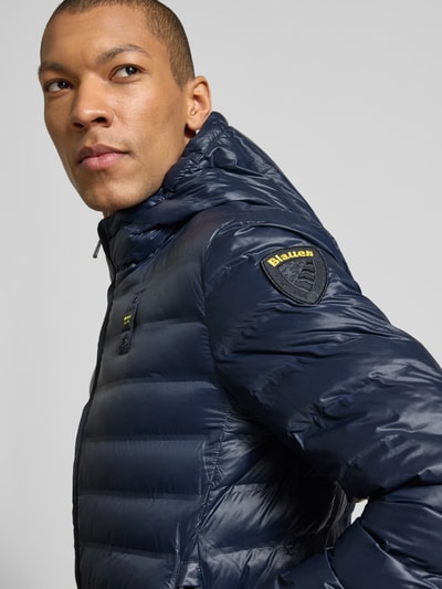 Männermodel trägt Steppjacke mit Zweiwege-Reißverschluss Modell 'Charles' von Blauer Usa in Dunkelblau mit Label-Detail, Nahaufnahme Oberkörper. Regular Fit, mit Kapuze, Rückenlänge 65 cm, Schulterbreite 47 cm, Brustweite 55 cm, Ärmellänge 65 cm.