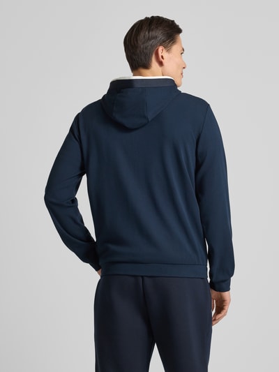 Męski model nosi Bluza rozpinana z logo i zamkiem błyskawicznym dwustronnym od EA7 Emporio Armani, mieszanka wiskozy, krój regular fit, kaptur, wpuszczane kieszenie, kolor granatowy, długość tyłu 68 cm, długość rękawa 68 cm, widok całej sylwetki od tyłu.