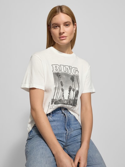 Frauenmodel trägt T-Shirt mit Statement-Print von Anine Bing aus Reine Baumwolle, taillierte Passform, Rundhalsausschnitt, Motiv-Print, Farbtöne in Weiß und Grau, weiche Textur, Nahaufnahme Oberkörper.