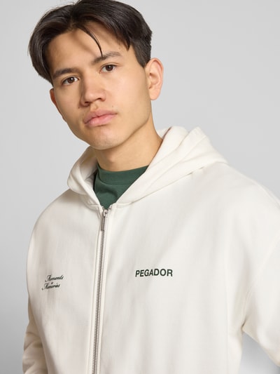 Männermodel trägt Oversized Sweatjacke mit Label-Print Modell 'Rochefort' von Pegador aus Baumwoll-Mix in Off-White, Nahaufnahme Oberkörper.
