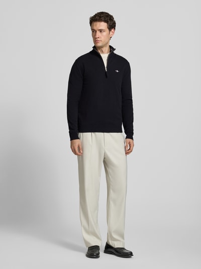 Männermodel trägt Pullover aus reiner Wolle von GANT in Schwarz, Feinstrick mit Troyer-Kragen und Reißverschluss, Ganzkörperansicht von vorne.