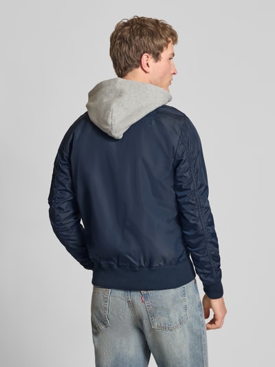 Männermodel trägt dunkelblauen Blouson mit Logo-Anhänger Modell 'MA-1 TT' von Alpha Industries, Sweat-Kapuze, elastische Abschlüsse, Ganzkörperansicht Rückansicht. Kontext: Herren Blouson, lockere Passform, durchgehender Reißverschluss, Armtasche, Label-Patch, Rückenlänge 69 cm, Schulterbreite 46 cm, Brustweite 54 cm, Ärmellänge 67 cm.