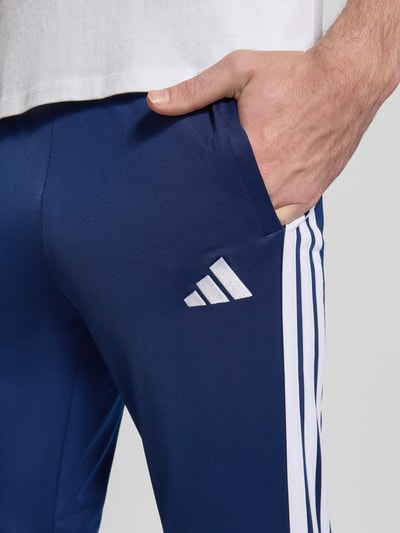 Männermodel trägt 'Trainingsanzug mit elastischen Abschlüssen' von adidas Sportswear, dunkelblau mit weißen Streifen, Detailaufnahme Oberschenkel. Szerokość w rozmiarze M: 36 cm, długość wewnętrzna nogawki: 75 cm, elastyczny pas, wpuszczane kieszenie.