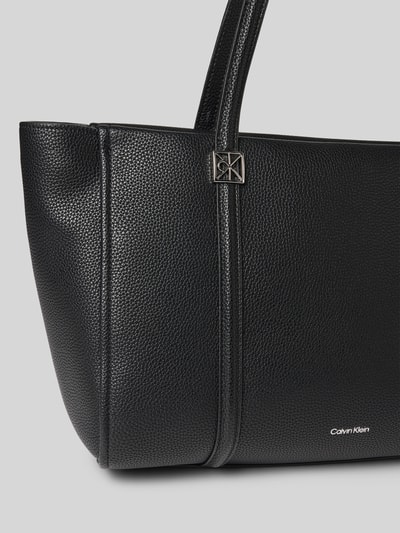 Produktfoto, Shopper mit Label-Detail von Calvin Klein, czarny kolor, faktura skóry, srebrne logo, Szerokość w rozmiarze 1: 47 cm, długość: 26 cm, głębokość: 16 cm, długość paska na ramię: 63 cm, główna komora na suwak, kieszeń wewnętrzna na suwak, uchwyty do noszenia.
