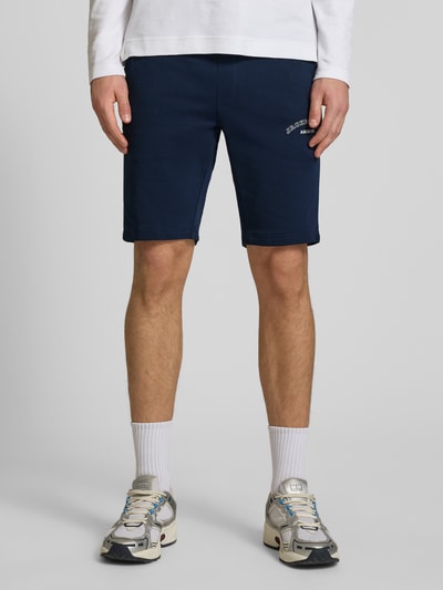 Männermodel trägt dunkelblaue Shorts mit Logo-Print Modell 'GORDON' von Jack & Jones aus Baumwoll-Mix, Relaxed Fit, Ganzkörperansicht von vorne.