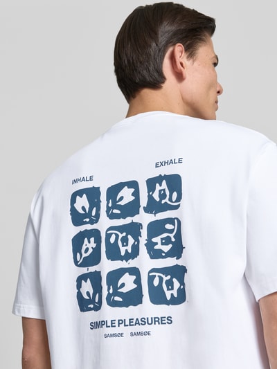 Męski model nosi T-Shirt mit Label-Print Modell 'COHEN' od Samsøe Samsøe w białym kolorze z niebieskim nadrukiem graficznym, czysta bawełna organiczna, krój relaxed fit, widok całej sylwetki z tyłu.